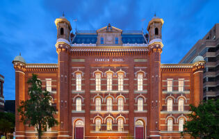 The front facade of the Planet Word building at night. It is a historic red brick building with decorative windows, cupolas, and a mansard roof.